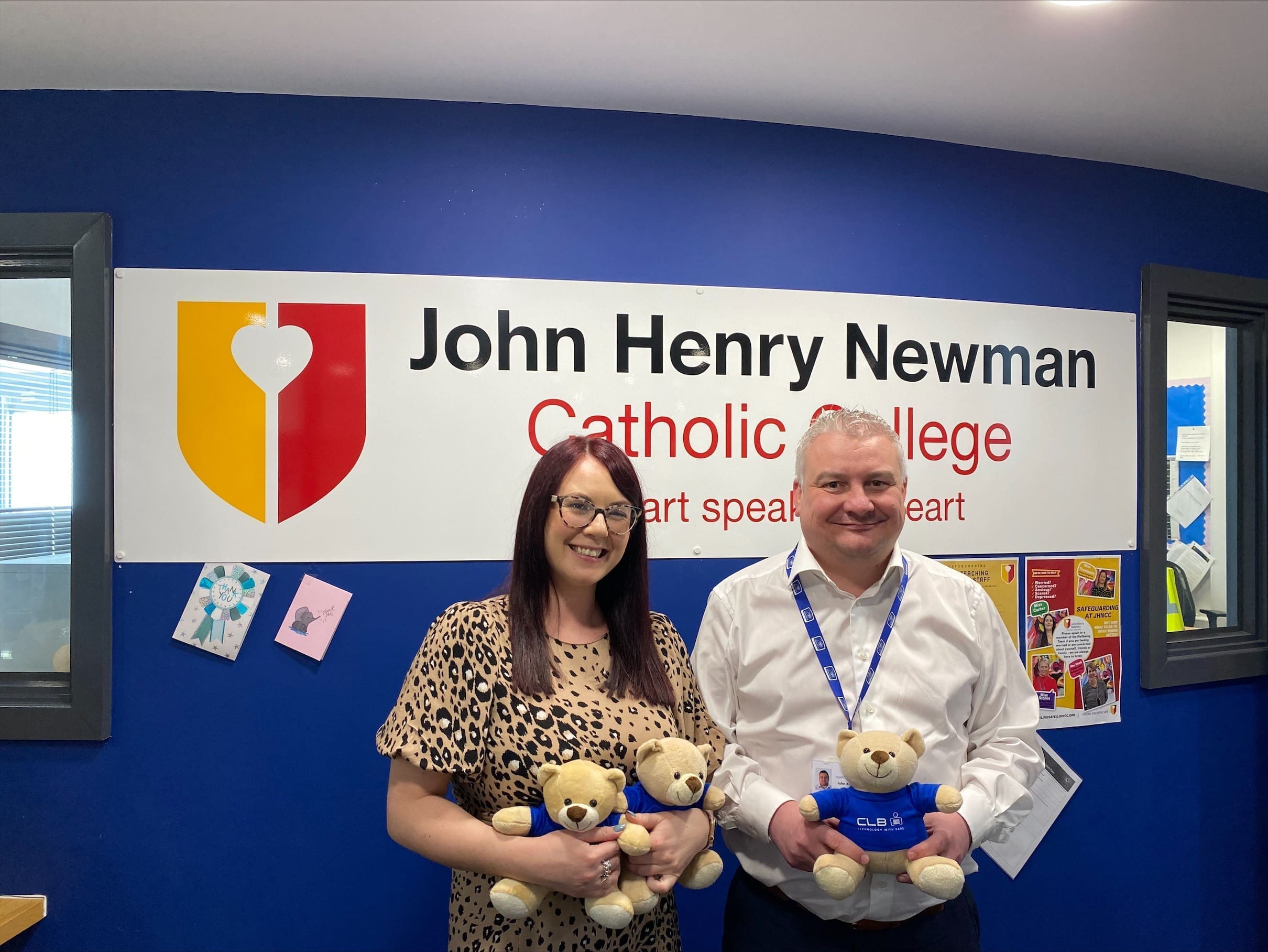 Two people hold teddy bears and smile in front of a sign that says John Henry Newman Catholic College